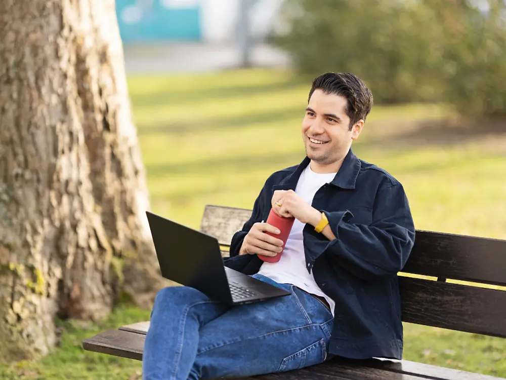 En Henkel-anställd sitter på en bänk i en park och arbetar på sin bärbara dator. 