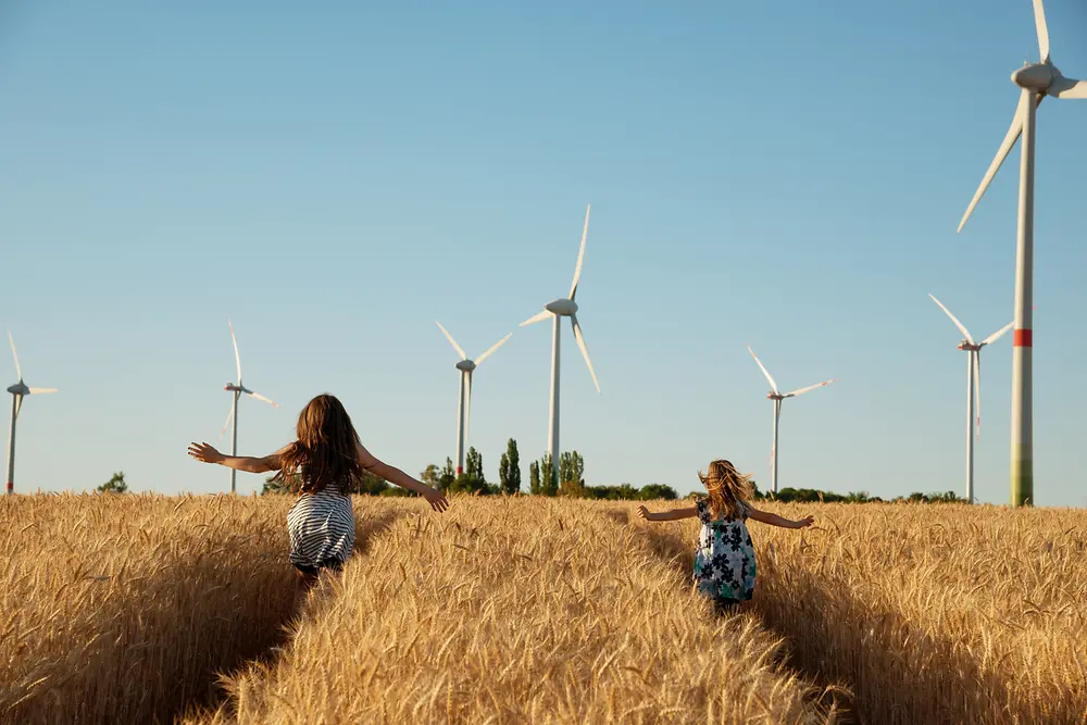 Flickor som springer genom en äng mot vindkraftverk. 