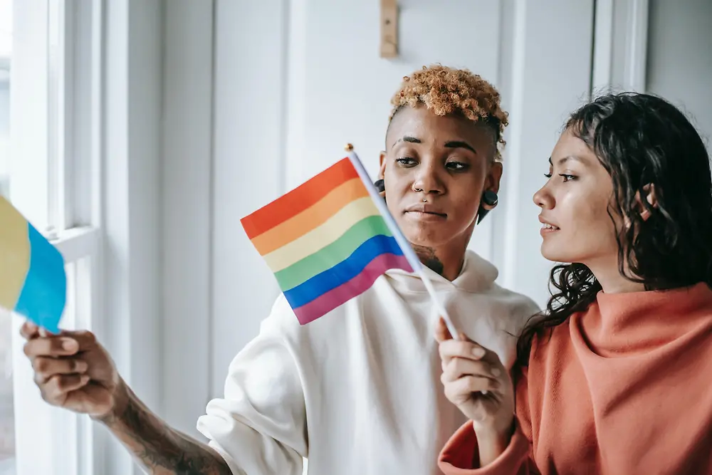 Två kvinnor pratar med varandra samtidigt som de håller en LGBTQ+ flagga.