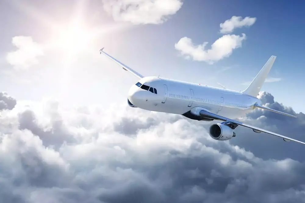 Ett vitt passagerarflygplan som flyger över mjuka moln på en ljus himmel, med solljus som skiner från övre vänstra hörnet.