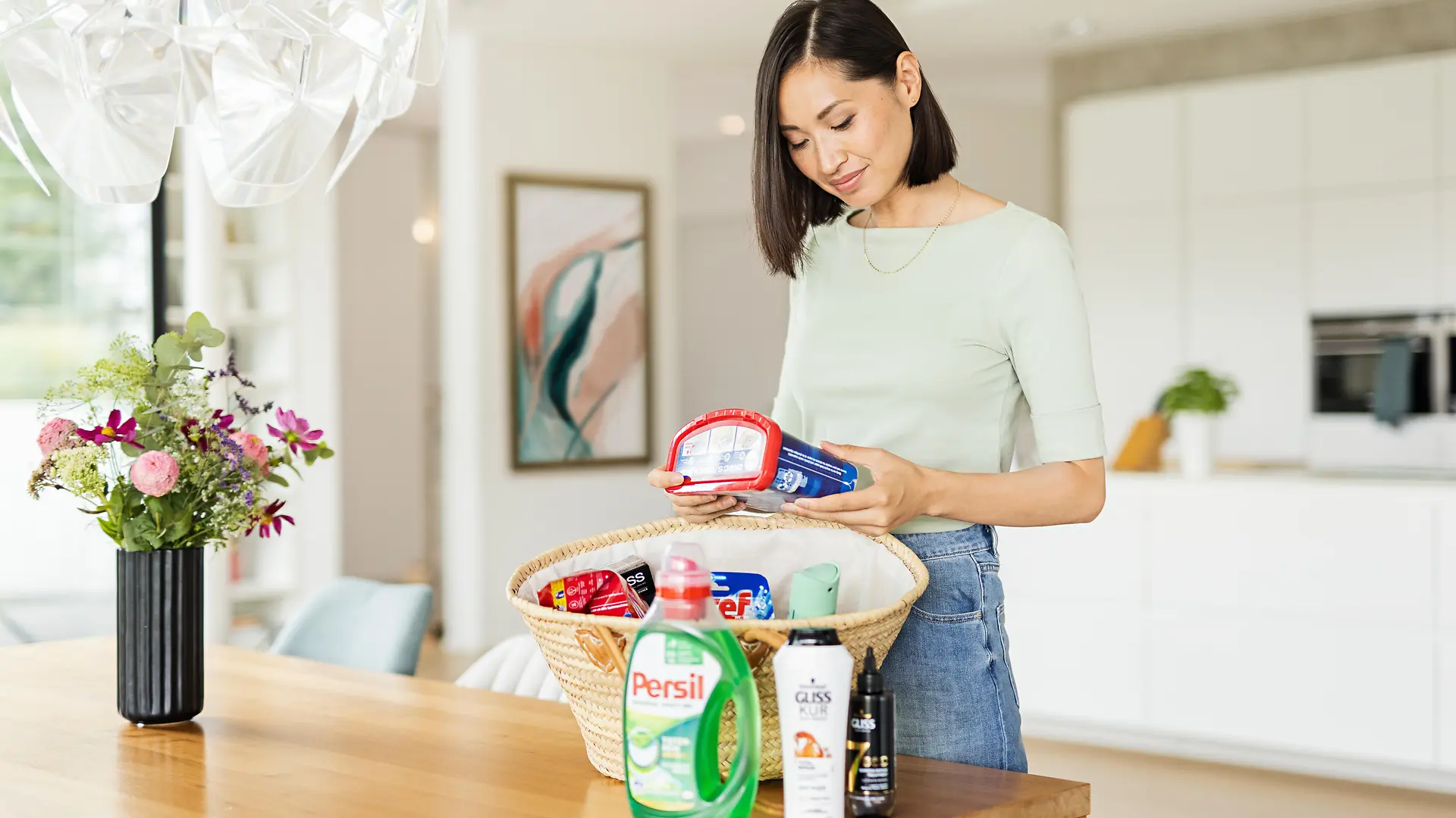 Kvinnor med en korg stående på ett bord som är fullt av Henkel-produkter som: Persil, Gliss, Bref, Somat, taft. och mer.