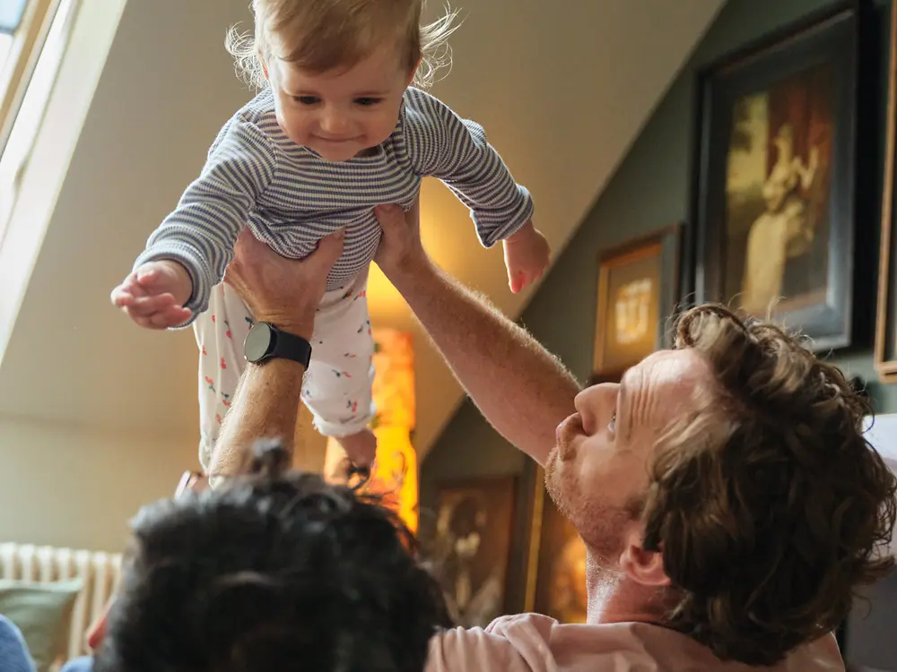 Två pappor som håller sitt barn och ler hemma, vilket symboliserar inkludering och Henkels familjevänliga policy för föräldraledighet.