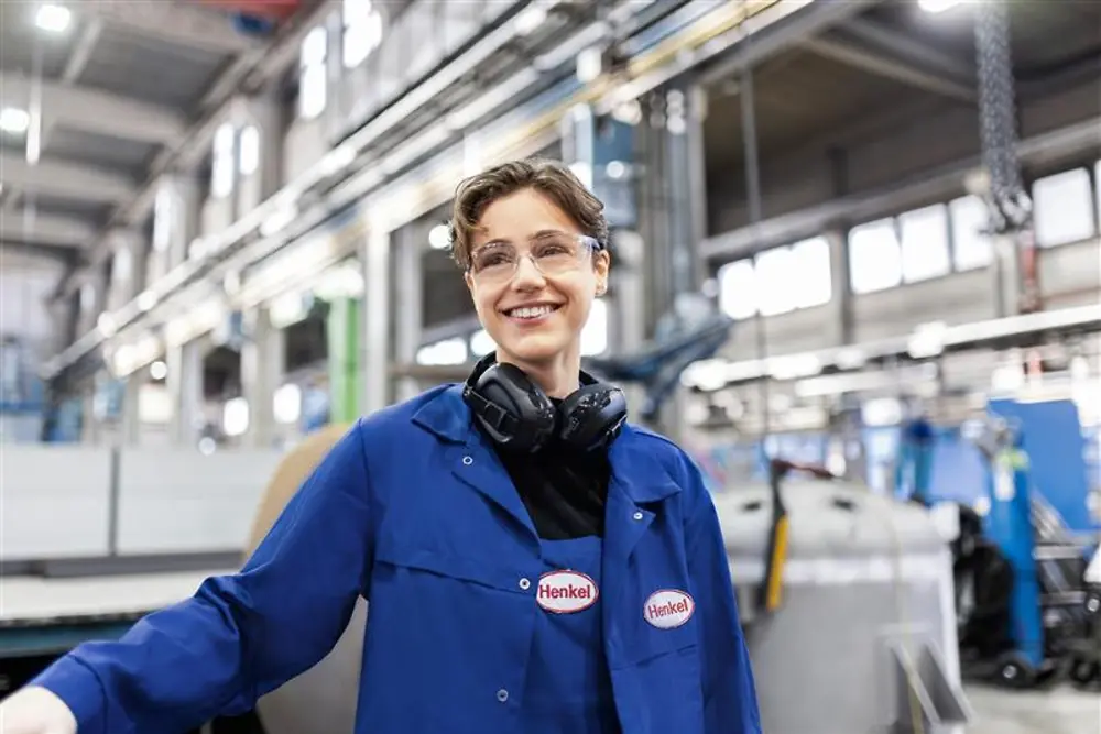 En leende Henkel-anställd klädd i en blå arbetsrock och med skyddshörlurar, som står i en industriell produktionshall och symboliserar expertis och innovation inom tillverkning.