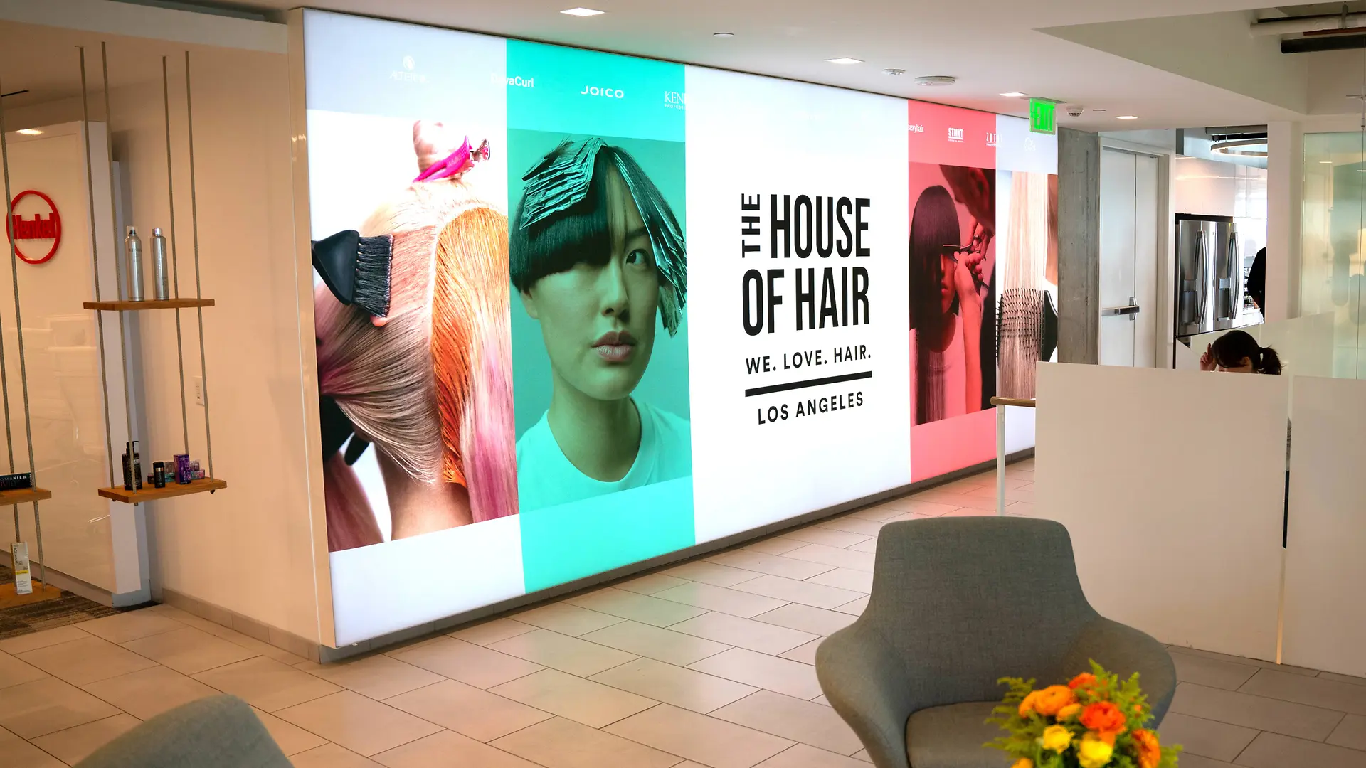 En modern lobby med en ljus väggdisplay med stora paneler med färgglada grafikmotiv på hår och texten ”THE HOUSE OF HAIR – WE. LOVE. HAIR. – LOS ANGELES.” Utrymmet har ljusgrå fåtöljer, ett litet bord med en livfull blomsterarrangemang och en ren, minimalistisk design med kakelgolv och vita skiljeväggar.