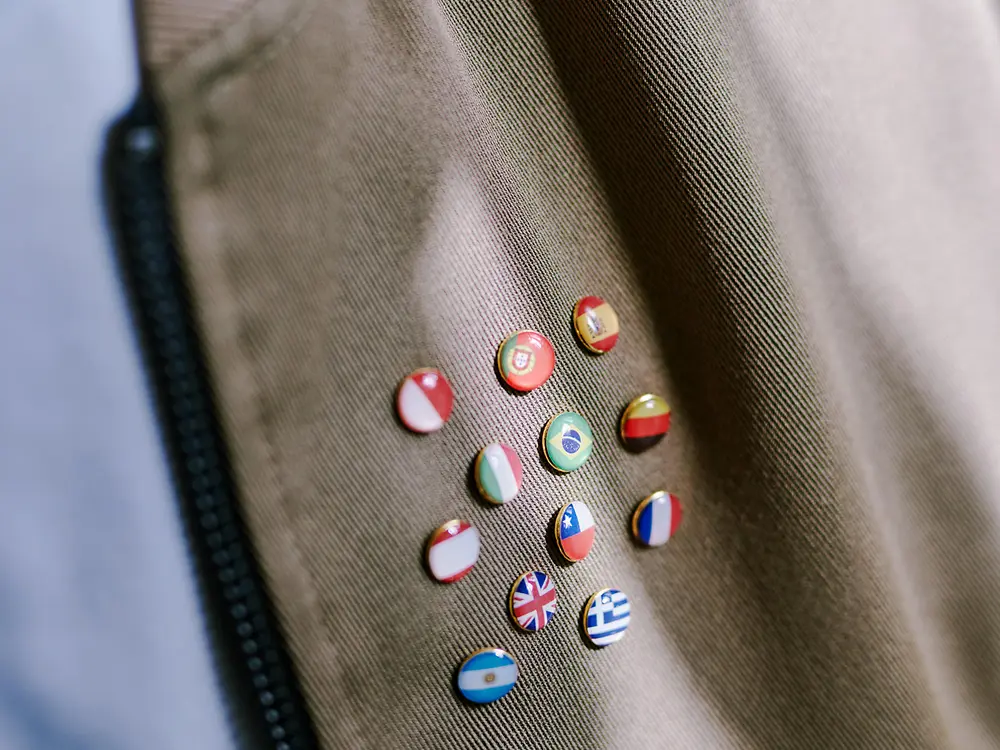 Närbild av ett brunt tyg med en dragkedja till vänster och flera små runda nålar som visar olika nationsflaggor, vilket symboliserar internationell mångfald.