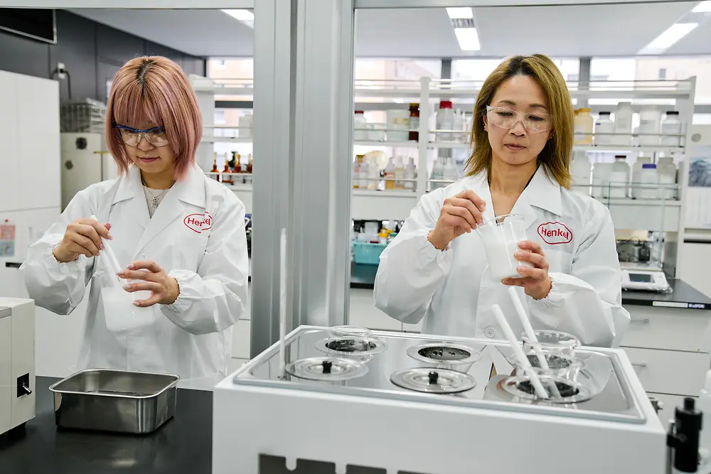 Två personer som bär vita Henkel-labbrockar arbetar i ett modernt laboratorium. De hanterar vetenskapliga instrument och behållare på en arbetsbänk, med diverse laboratorieutrustning och hyllor med förnödenheter synliga i bakgrunden.