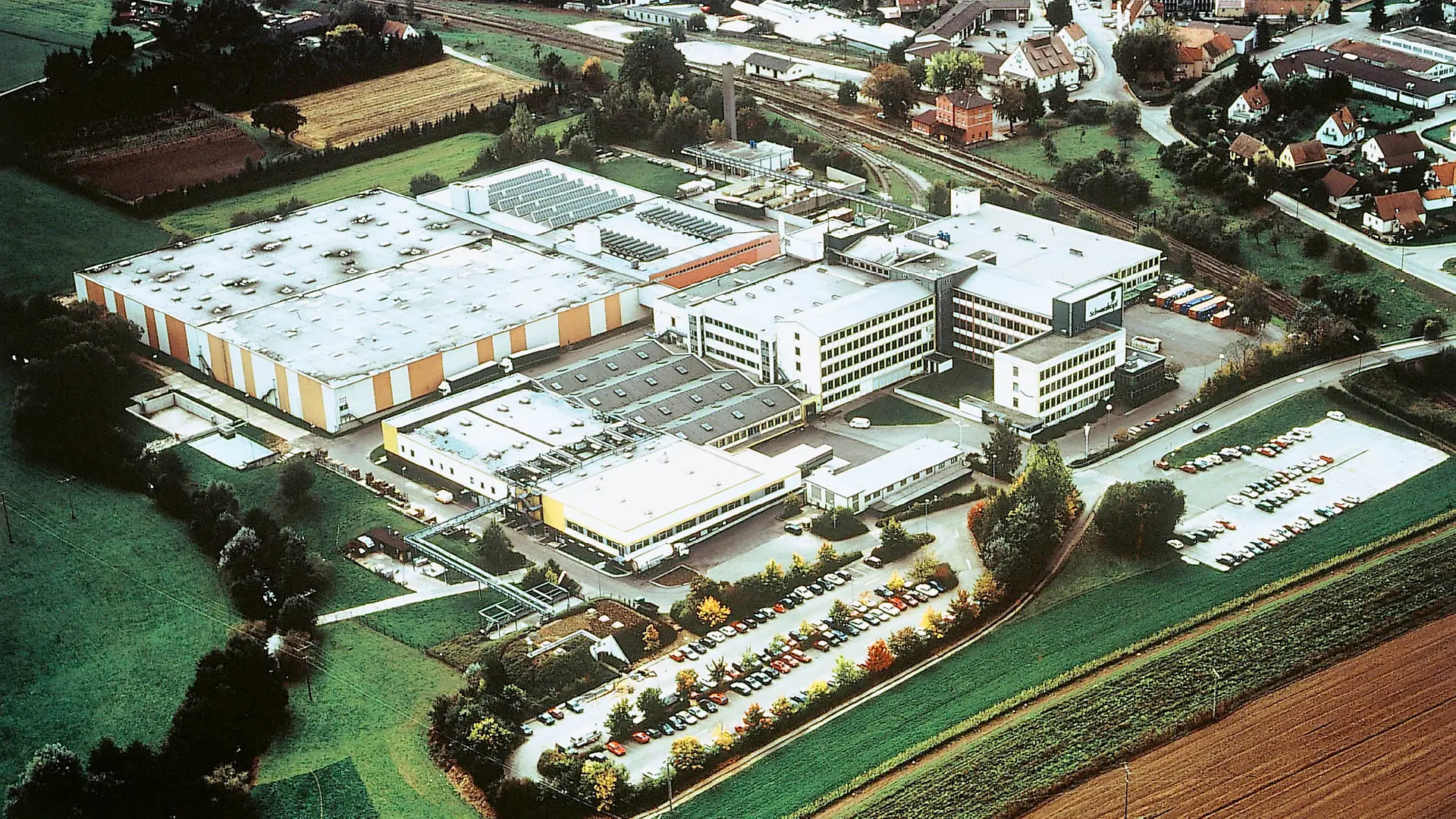 En flygbild över Schwarzkopf-fabriken i Wassertrüdingen 1995, som visar flera produktionsbyggnader och hallar samt en stor parkeringsplats full av bilar.
